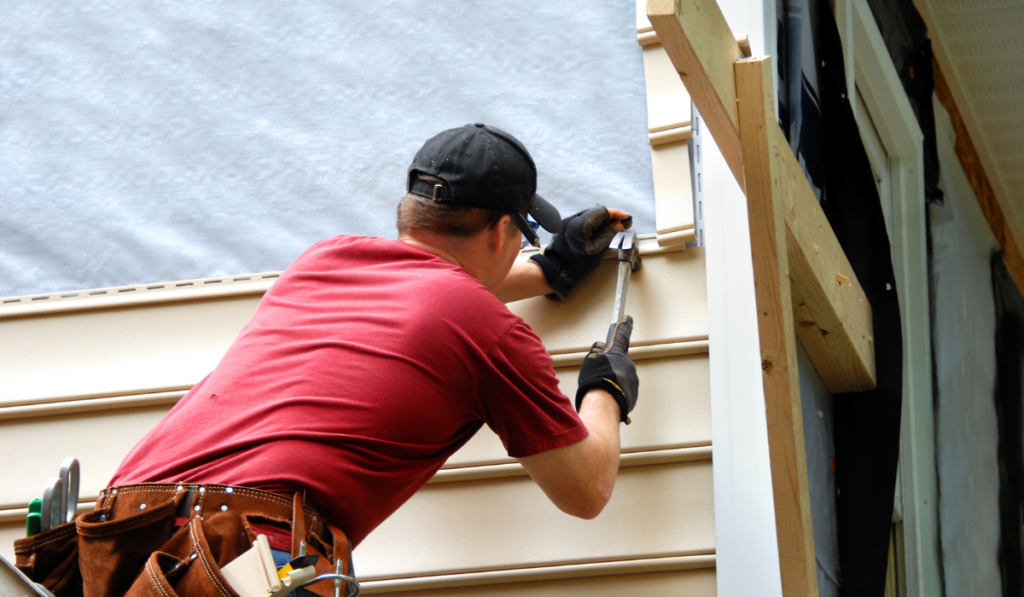 Young homeowner installs siding to his home. He is holding a hammer and wearing a tool belt