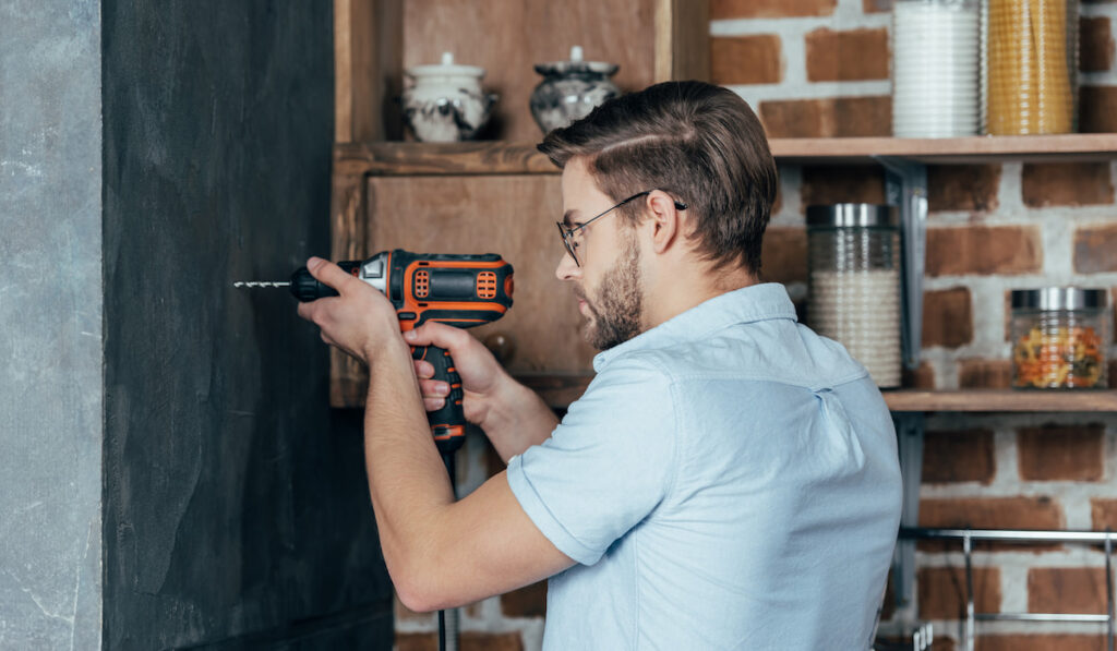 side view of young man drilling wall with electric drill at home
