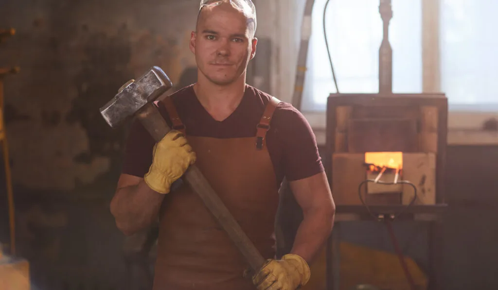 Portrait of worker in apron and protective gloves standing against furnace and holding sledgehammer