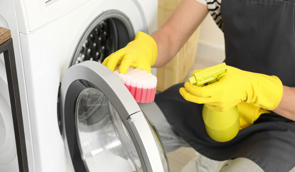 man cleaning washing machine using vinegar on spray bottle wearing yellow gloves
