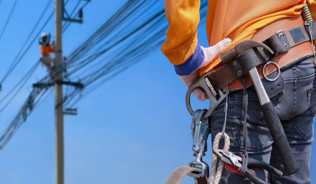 rear view of electrician with electrician hammer on his safety belt with blurred background of electrical workers team on power poles