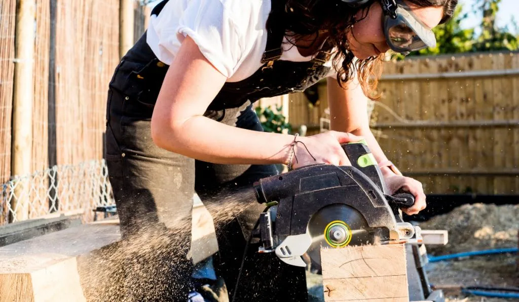 Woman wearing protective goggles and ear protectors holding circular saw