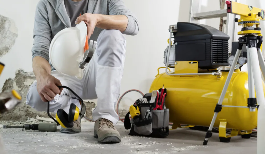 house renovation concept, construction worker holds in hands helmet and safety headphones, air compressor and construction tools in the background
